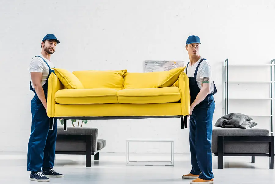 Two professional movers in blue overalls and caps carrying a bright yellow sofa across a bright modern room Furniture Removal Service and also Junk and Clutter Removal in Seattle WA